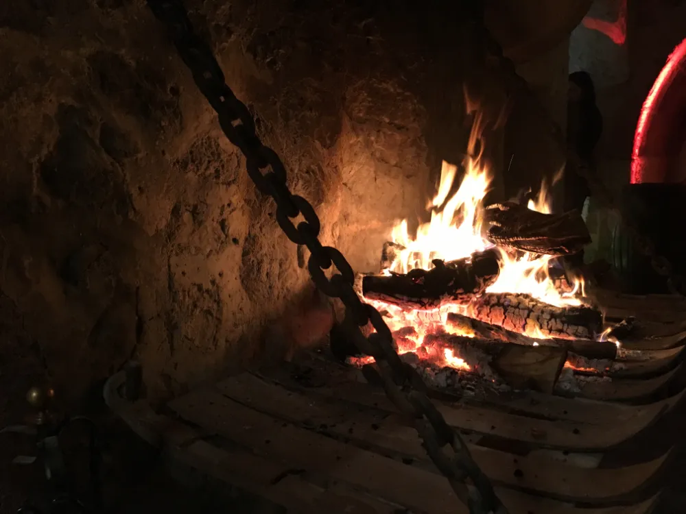 Roaring wood fire with glowing embers set against a rough stone wall, partially obscured by a heavy metal chain hanging in the foreground
