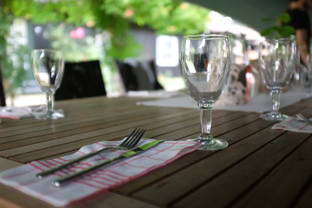 Close-up of a wooden outdoor dining table set with empty clear glasses and silverware resting on white and red grid-patterned napkins, with a blurred background of greenery