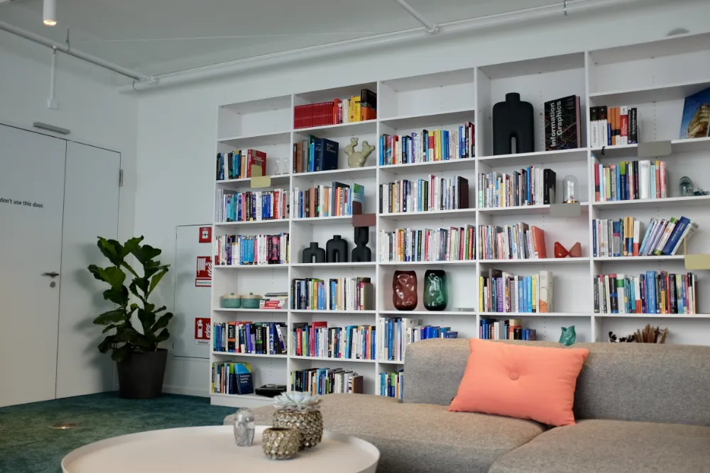 Bright office library corner featuring a large floor-to-ceiling white bookshelf filled with books and decorative objects, a grey sofa with a peach pillow, and a potted plant next to a white door